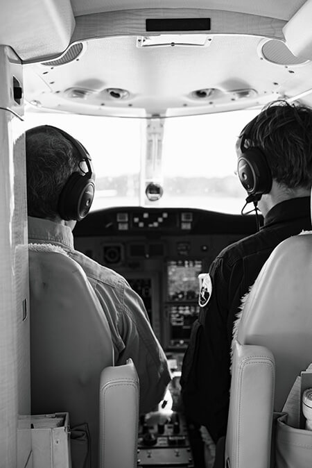 Two pilots wear headsets and face toward the front of the plane looking over the instrument panels in the cockpit